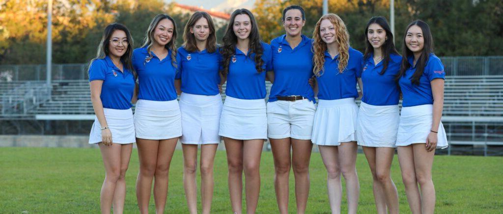 六名女子高尔夫队员在球场上站成一排. 他们穿蓝色衬衫和白色短裤和裙子作为制服.